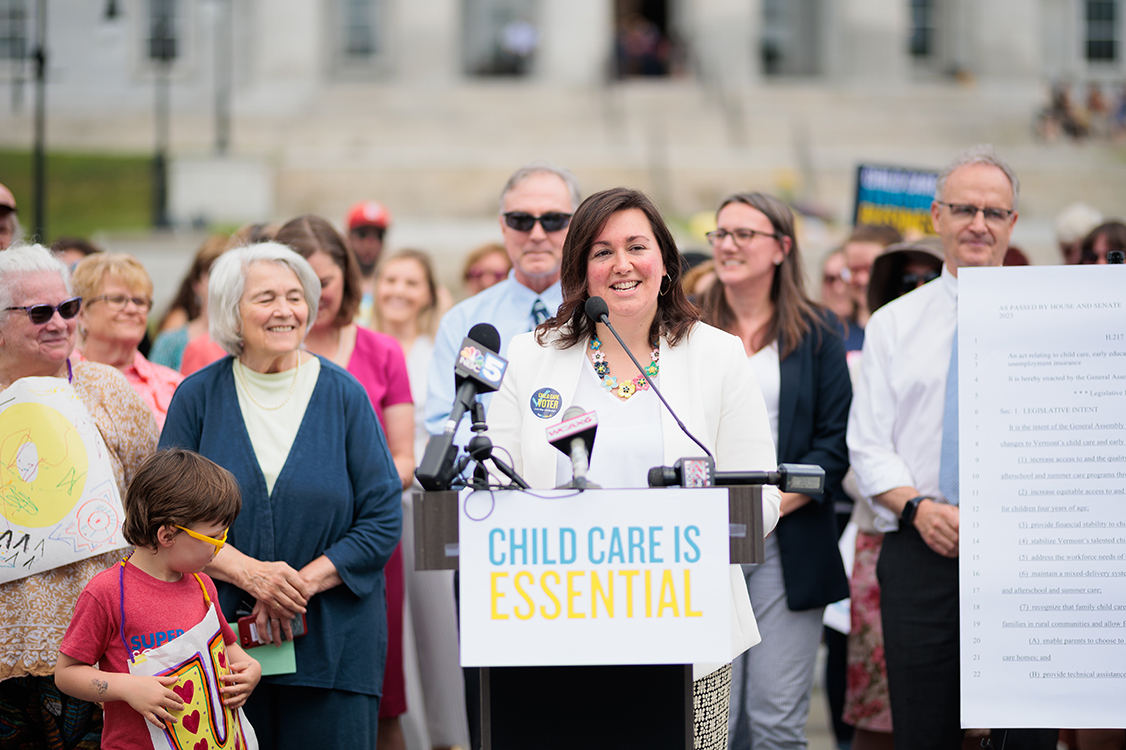 Aly Richards standing at a podium, speaking at a rally. The sign on the podium says "Child Care is Essential." There are many people standing behind her in support.