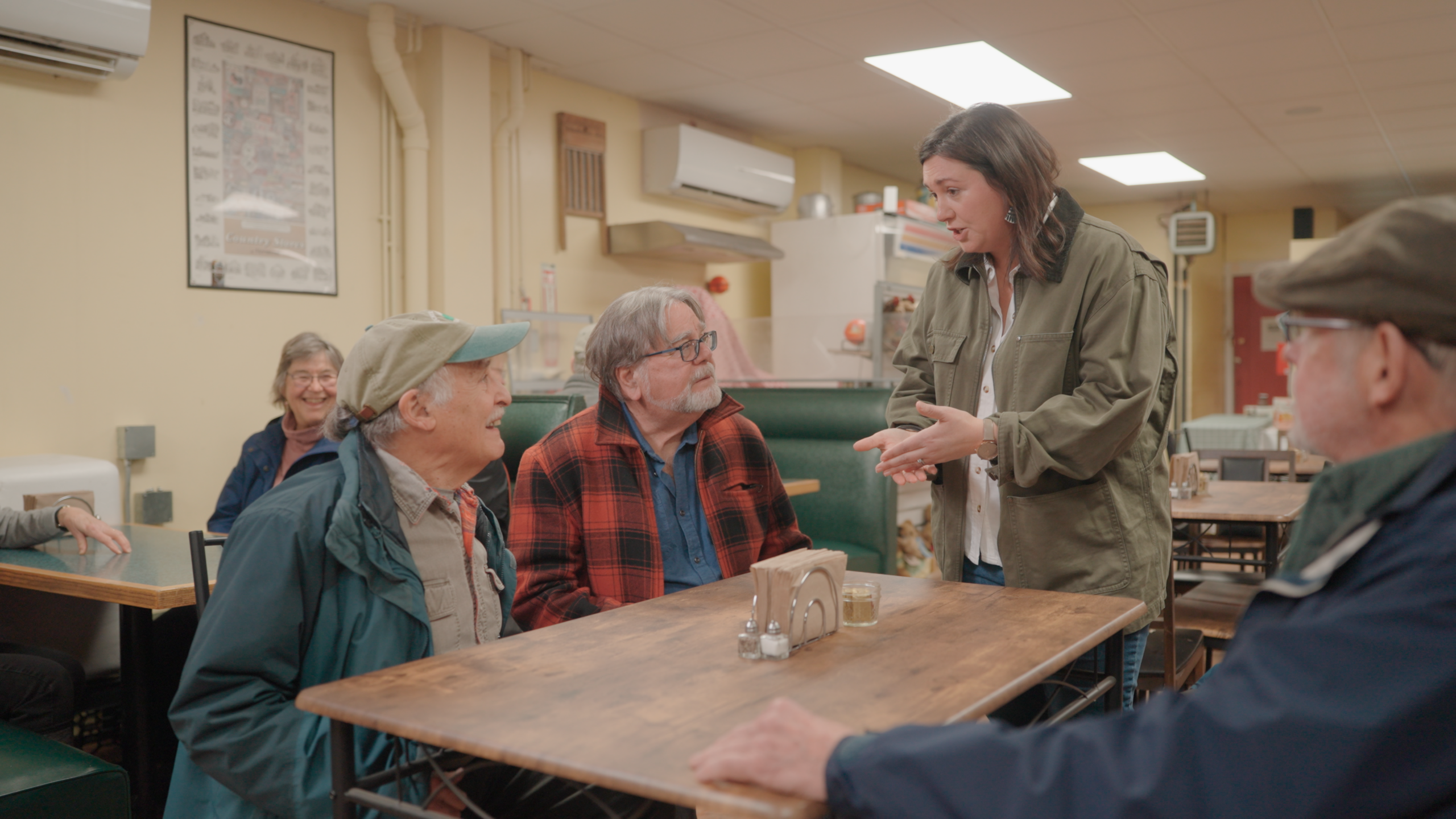 Aly Richards is standing by a table in a diner, talking to three older men.