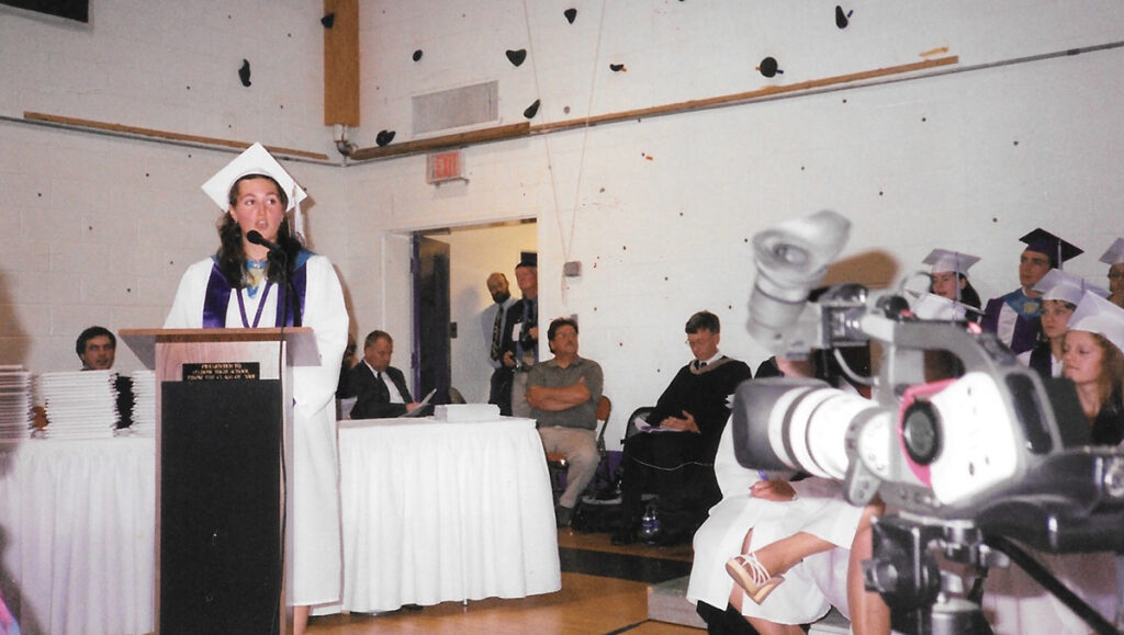 Aly Richards, as a teenager, graduating from high school. She is wearing a cap and gown and standing at a podium talking.