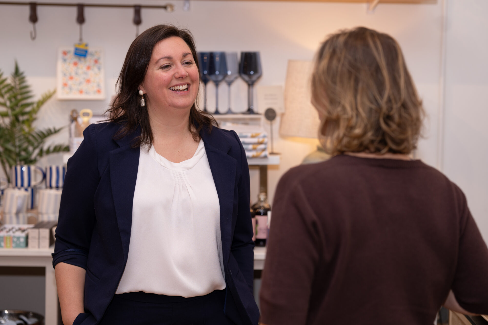 Aly Richards, dressed in a white shirt with a blue blazer, is talking to a woman in a brown sweater. The camera is positioned behind the woman she is talking to, so we only see the back of her head. They are standing in a shop.